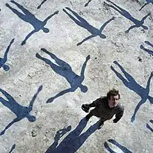 A man standing on a sidewalk with many people's shadows flying over him in the background