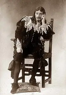 white man in 17th-century costume with long wig and small moustache and pointed beard, sitting pensively in a chair