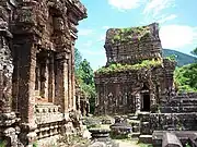 Ruins of buildings of red stone with niches and sculptures. The roof of one of the structures is partially covered in grass.