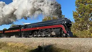 A black streamlined steam locomotive with a 4-8-4 wheel arrangement (four leading wheels, eight driving wheels, and four trailing wheels) and its tender, pulling an extra tender with the in the background