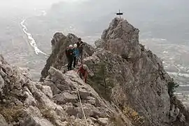Hikers crossing a tricky rocky ridge with a small metal cross in the background and a settlement in the background.