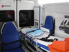 Interior view of a KTW: transport chair (left) gurney (middle) paramedic's chair(right). One can also see the O2 – bottles (cabinet in the rear) and the medical ventilator (right foreground)