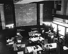 An electronic simulation room at the Naval War College during a 1958 wargame: against the far wall, a large map shows the outline of landmasses and some firing solutions.  Suited men sit at desks on the floor, papers in front of them, most staring up at the map.  Against the right wall, uniformed ensigns plot ship locations on (washed-out) screens.