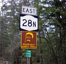 Three signs, from high to low: A rectangular sign that reads "EAST", a New York state highway sign, and a brown-and-yellow trail marker