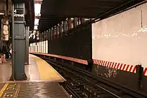 View of the southbound local platform of the 14th Street–Union Square station on the IRT Lexington Avenue Line. Behind the wall and the black bars on the right there is an abandoned side platform, whose edge is still visible. The mezzanine is right above. The red and white stripe indicates that there is no clearance between trains and the wall. In the foreground one can see the gap fillers that are automatically operated via proximity sensors. The writing on the floor says "stand clear".