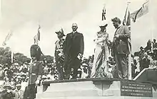 Five people on stone platform, from left: Man with mustache in military dress uniform and bushy tall black hat with chinstrap; man in military uniform with many medals and old fashioned naval officer's hat; man with white hair in suit with long dark coat and white collar; woman in Victorian dress with white hat; man with beard and wild hair in suit. Crowd and flags in background.