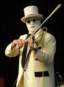 A black background. In the foreground, a man in a white suit with black lapels and black pocket flap, wearing a white top-hat and glasses with a thick circular frame plays an electric violin which is nestled under his left cheek. His face is covered by a layer of surgical bandages, with only a portion of his forehead exposed and a slit at the mouth. A yellow wire hangs from the top of the violin at his left shoulder.