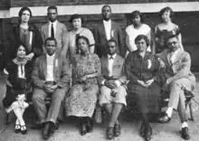 Twelve African-American adults, seven women and five men, posed outdoors for a group photograph, one row standing, one row seated.