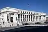 A white stone, classically designed, 4 story building. Doors are located at the corners of the building, on the second story with a grand staircase up to it, and are flanked by two ionic columns.