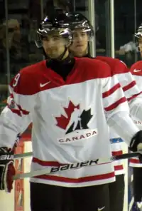 Hockey player in red and white Canada uniform. He has a half-smile on his face and holds his stick in his hands.