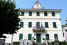 A color photo of a three story building with shuttered windows, three flags, and a clock tower.