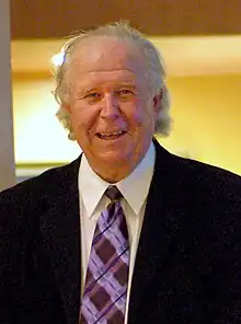 A man with disheveled white hair wearing a black suit jacket and purple tie looks straight ahead and smiles.