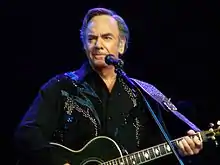 A Caucasian male singing into a microphone. The male is wearing a black suit with a beaded design on the front and is holding a guitar with both his hands.