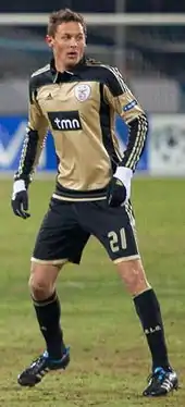 A young man on a football pitch during a match. He is wearing a bronze jersey with black details, and black shorts, socks and gloves.