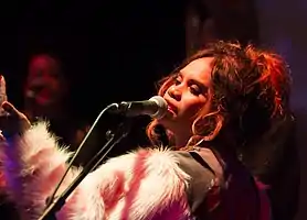 A 31-year-old woman is shown in upper body and left profile. She is singing into a microphone while raising her left arm in front of herself. She wears a khaki cloth top with pink and white woolly sleeves. Another person is blurred in the background.