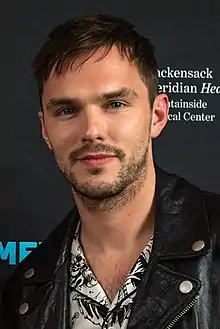 A young, Caucasian man with short, dark hair and facial stubble wearing a black shirt looks away from a microphone against a grey and blue background.