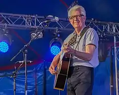 A white-haired man smiles as he strums his guitar on stage