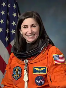 A female Caucasian astronaut wearing her orange mission suit with the American flag embroidered on her shoulder, and name tag and mission patches embroidered on her chest.