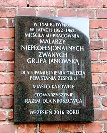 A black plaque with Polish text on a red brick background. The Polish text says that a group of "non-professional artists" operated in this building from 1952-1962, and the plaque commemorates the 70th anniversary of the founding of the group.