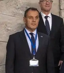 56-year-old man standing at bust length with dark jacket and tie, light blue shirt and electric blue badge holder lanyard