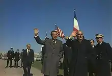 Two men in suits stand to the right, with uniformed military officers nearby. Both men are waving and smiling.