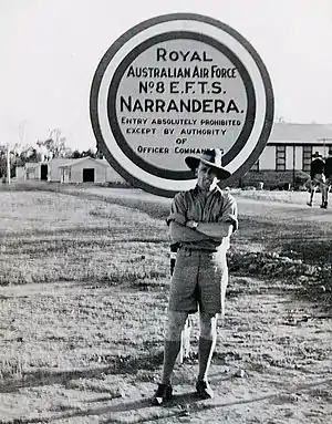 Informal portrait of uniformed man in slouch hat standing in front of sign reading "Royal Australian Air Force No. EFTS Narrandera"