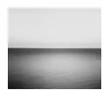 A black-and-white image of a still sea meeting the sky, with the horizon running through the centre. The sun's reflection is visible on the water.