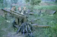 A damaged artillery piece sits in the foreground while a number of rifles resting against it. In the background a number of Caucasian soldiers stand in the background next to a hootchie.