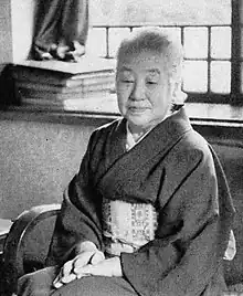An older Japanese woman, seated indoors, wearing a kimono, hands clasped in her lap