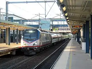 A silver train with blue and red accents hauled by an electric locomotive at a train station