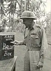 Three-quarter-length outdoor portrait of moustacioed man in light tropical military uniform with pith helmet and pilot's wings on left breast pocket, placing folded paper into a box marked "RAAF BALLOT BOX"