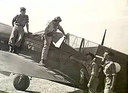 Pilot with goggles emerging from cockpit of single-engined monoplane which has the letters "VE" prominently displayed on its fuselage, in company with three other men