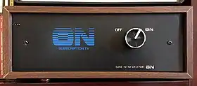 A front view of a wooden box with wood-veneer exterior and a black front. A blue "ON Subscription TV" logo, with the ON letters in a linear gradient, graces the front. A silver knob selects between two options: OFF and ON (stylized like the logo). Beneath the knob is the instruction "TUNE TV TO CH 3 FOR ON".