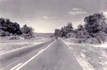 A black and white photograph shows a single paved road with a painted centreline. Vehicles can be seen driving the road in the distance. Trees abut against the right side of the road, while the left side features a wide landscaped clearing before the tree line.