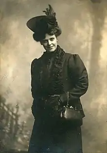 A white woman, standing and smiling, wearing a dark hat, coat, gloves, and holding a dark handbag over one arm.