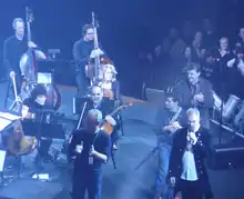 A small orchestra mostly compromising of the string section is playing on stage, with one person holding a microphone and the audience are on the topright of the picture.