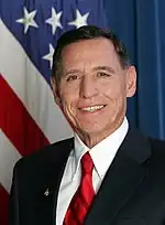 Head and shoulders of a man in his forties with close-cropped dark hair, dressed in a dark blue suit, a white shirt and a red striped tie. On his right lapel is the President’s "Call to Service" Award medal. Over his right shoulder the U.S. flag.