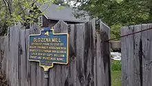 a picture of the marker, on a post like others but also attached to a fence through which you can see the stream that powered the mill