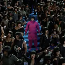 Oliver Tree as his character Cornelius Cummings, with a bob haircut and all-pink outfit, standing on a blue carpet surrounded by people all with the same bob haircut