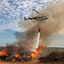 A helicopter passing over a bush fire, with a bucket slung below it. Water is being dropped on the fire from the bucket.