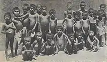  A group of 15 boys, 10 standing and five squatting. Most appear naked. All have prominent pot-bellies but ribs obviously showing, a common symptom of malnutrition.