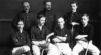 One row of four men seated with three men standing behind. Several have hockey sticks
