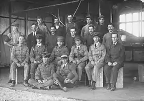 Group portrait of nineteen men, including six in military uniforms with peaked caps, in front of a biplane in a hangar