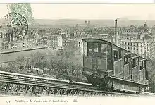 A sepia postcard taken at an angle to the track showing one of the first funicular's cabins. Its stepped compartments are clearly shown. In the background is a panorama of Paris