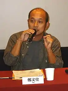 Cheng speaking into a microphone while sitting behind a red table.