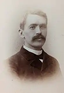 Black and white photograph of a man with a large moustache wearing a suit with a velvet collar and a shirt with a standing clergyman's collar.