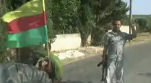 A flag with horizontal bars of yellow red and green attached to a military vehicle (left) beside a Kurdish YPG fighter holding an AK-47. (right)