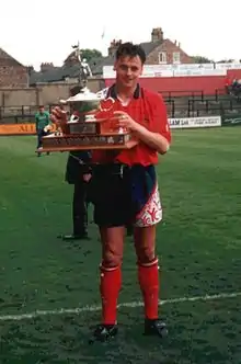 A man standing on a grass football pitch, wearing a red shirt, black shorts and red socks, and holding a trophy.