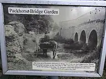 A sign showing a picture of the Packhorse Bridge in 1906 alongside information. The text reads as follows: This is the site of Kentford's old Packhorse Bridge, one of only two such bridges built in Suffolk. The photograph was taken on New Year's day in 1906. Since then, sadly, all that remains is the wall of the road leading to the bridge. The final parts of the bridge disappeared in the 1970's. Enjoy the bench and imagine times past!
