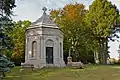 Almost white octagonal stone mausoleum with pointed roof. Intricate design wraps around the building just below the start of the roof. Door is aged metal.